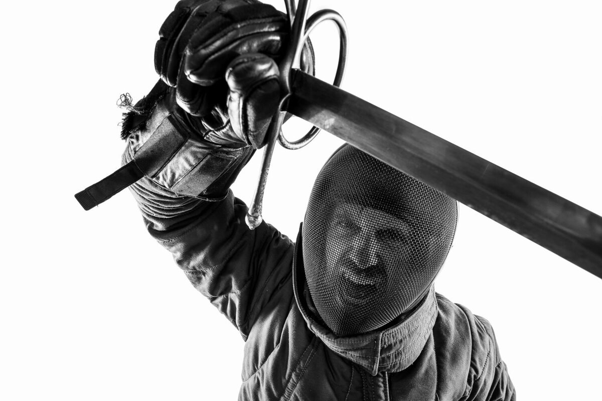 Dramatic editorial-style photo of a fencer in full gear raising a sword toward the camera.
