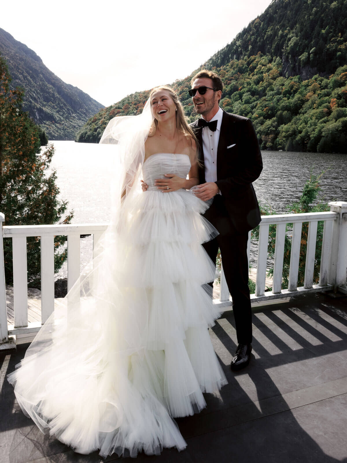 The bride and the groom are smiling and standing on a terrace, overlooking waters and mountains, at The Ausable Club, New York.