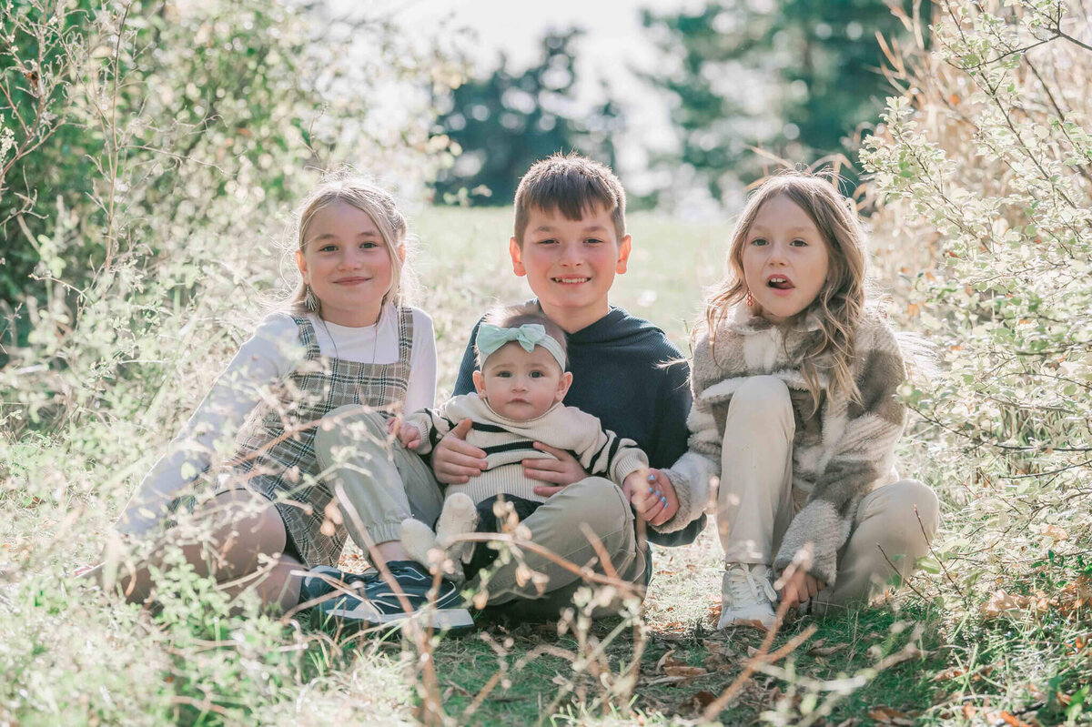 The four kids sitting on the grass in an opening of trees. 