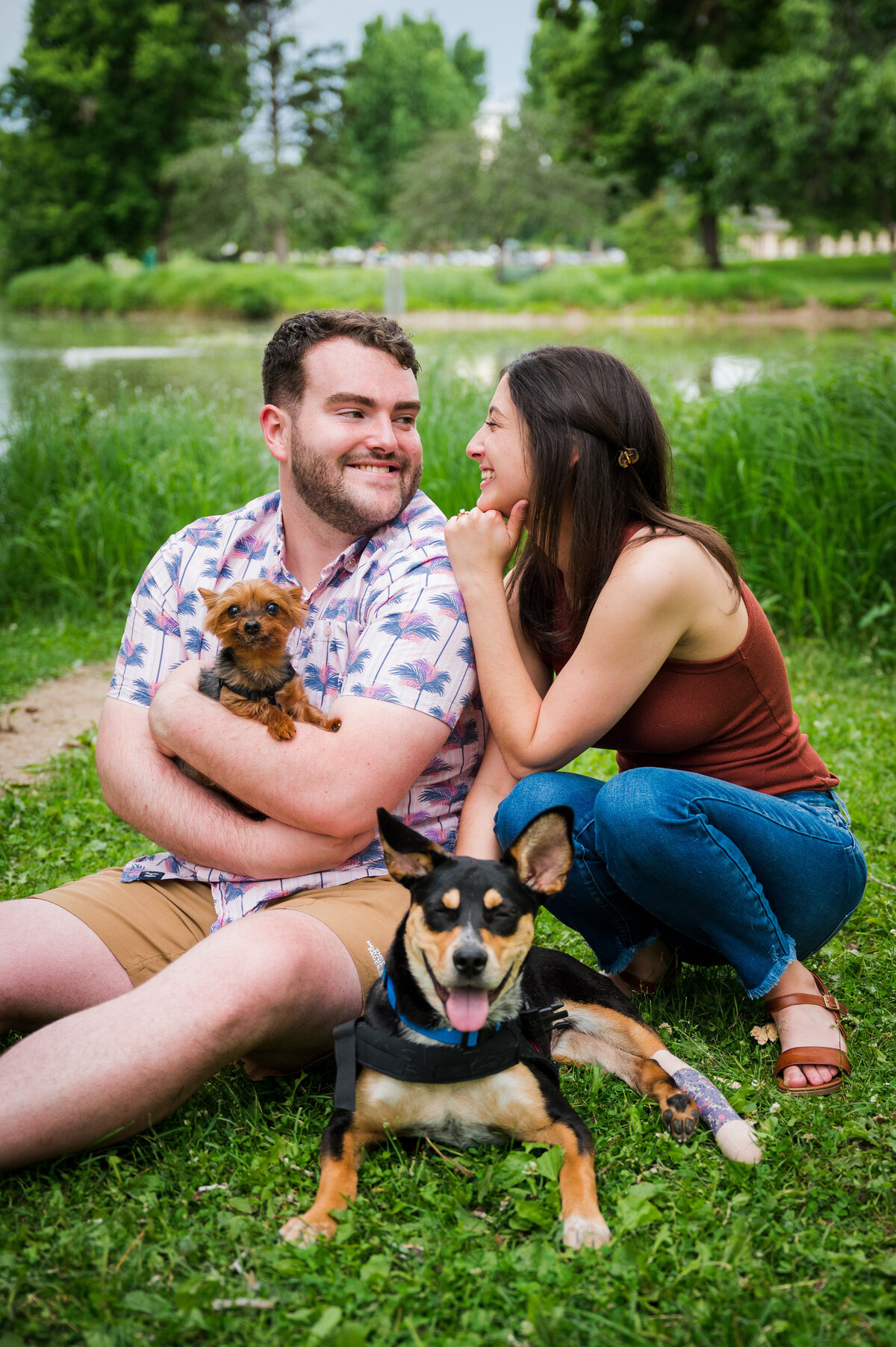 A man and woman sit in the grass cuddling up to each other. One of their dogs sits in front of them and the man holds the other.