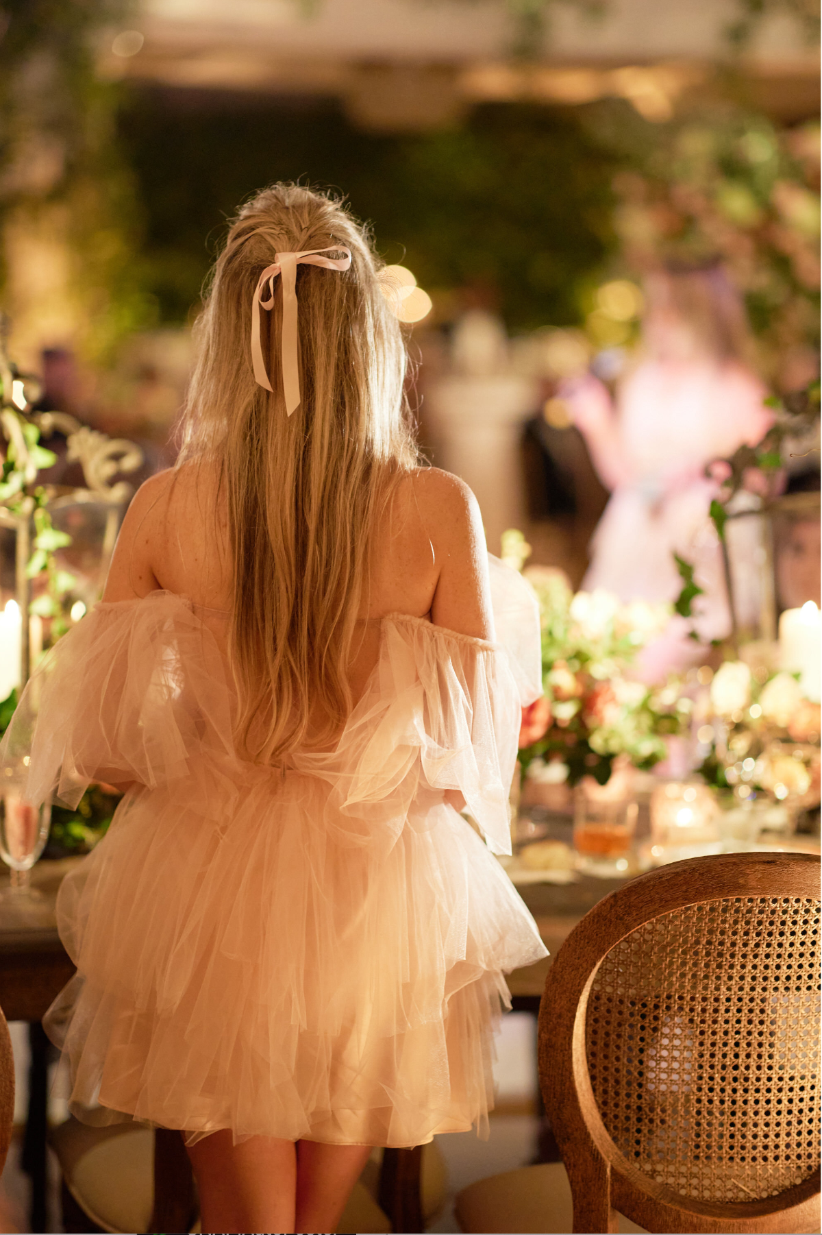 The photo was taken at the luxurious Willard Intercontinental in Washington D.C., known as one of the top wedding venues and hotels in the city. It was captured by photographer Shawn Connell. In addition, Clayton Films, known for their expertise as videographers, also contributed to the event. In the image, a woman with long blonde hair, adorned with a white bow, stands with her back to the camera. She is wearing a soft, off-shoulder, layered tulle dress. The setting is warmly lit, with elegant decor that includes candles and flowers, creating an atmosphere that suggests a formal or festive event.