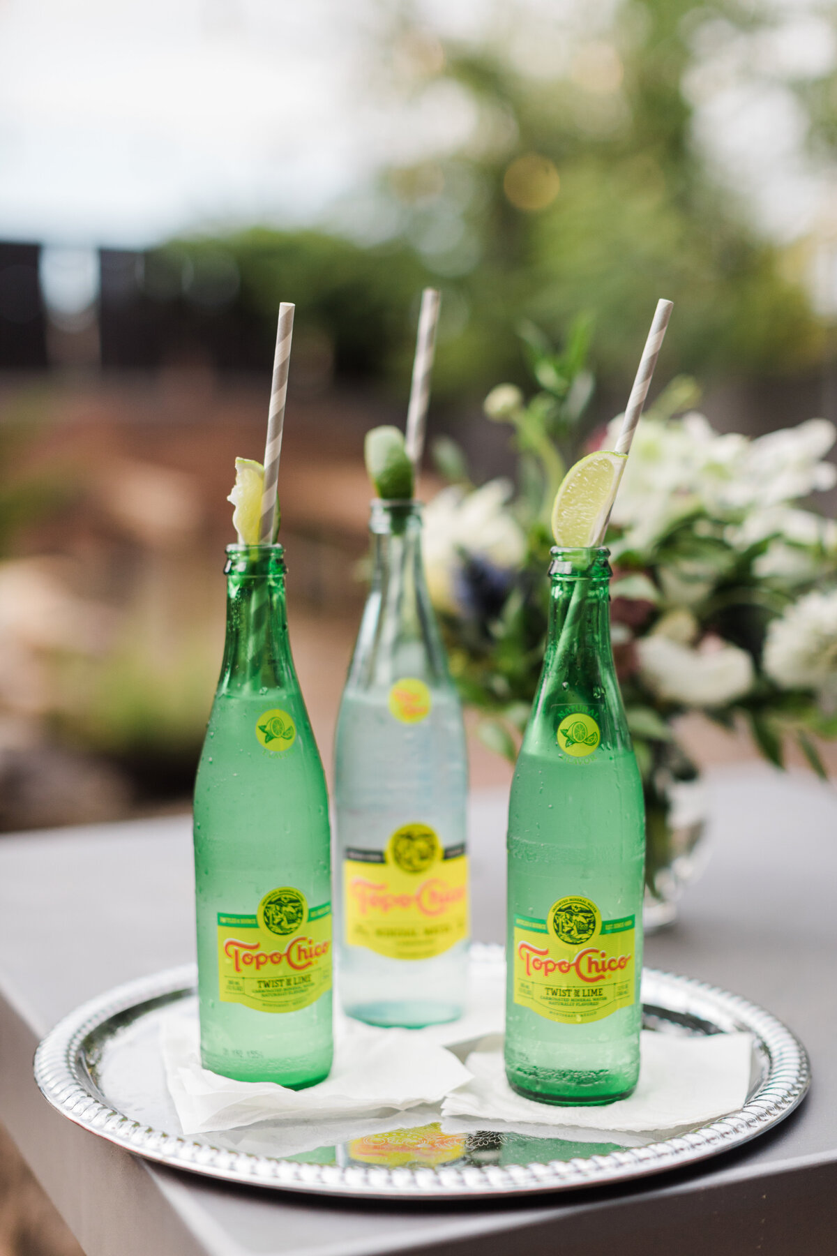 Detail photo of three bottles of Topo Chico on a silver platter during a wedding reception at Artspace111 in Fort Worth, Texas. Each bottle is resting on a napkin and has a lime wedge and straw coming out the top. A bouquet of flowers can be seen out of focus in the background.