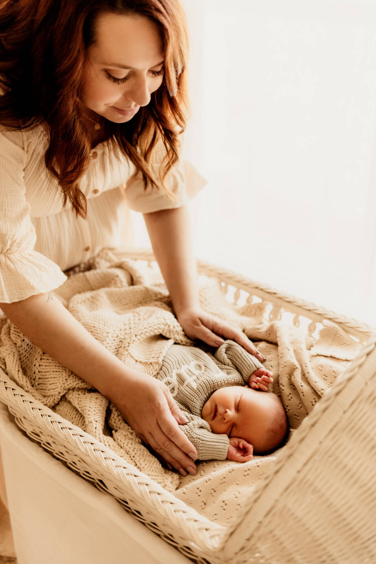 Mother wearing a cream dress tends to her newborn baby boy who is sleeping in a bassinet with his hands near his face.