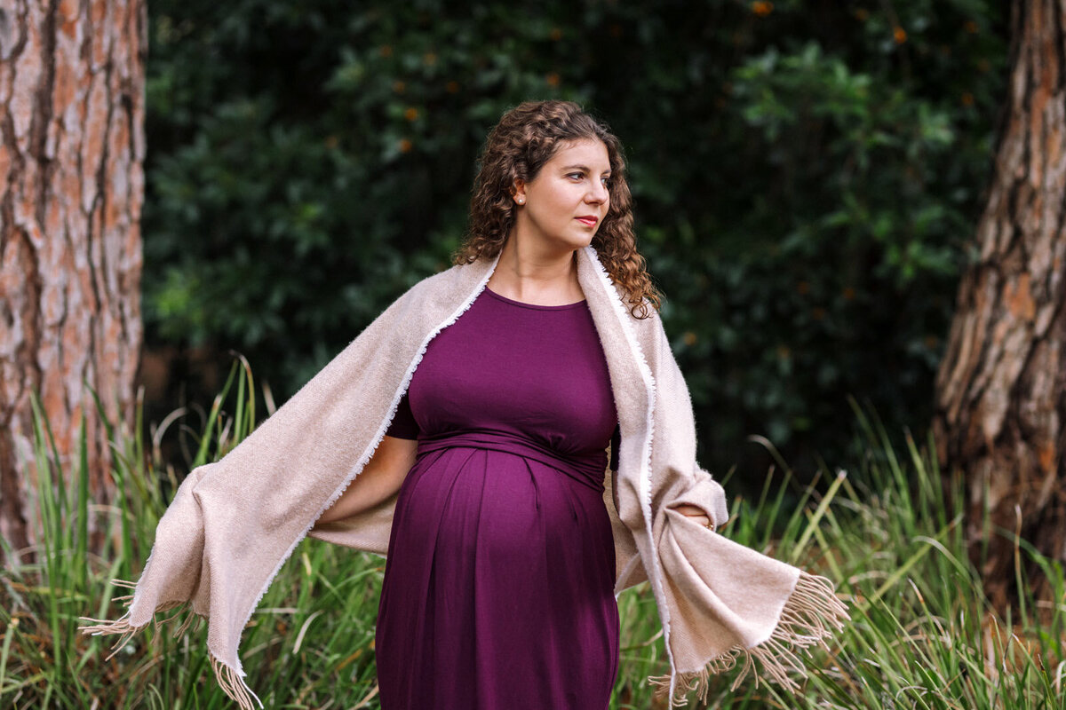 pregnant woman in purple  dress standing between trees and moving her scarf around