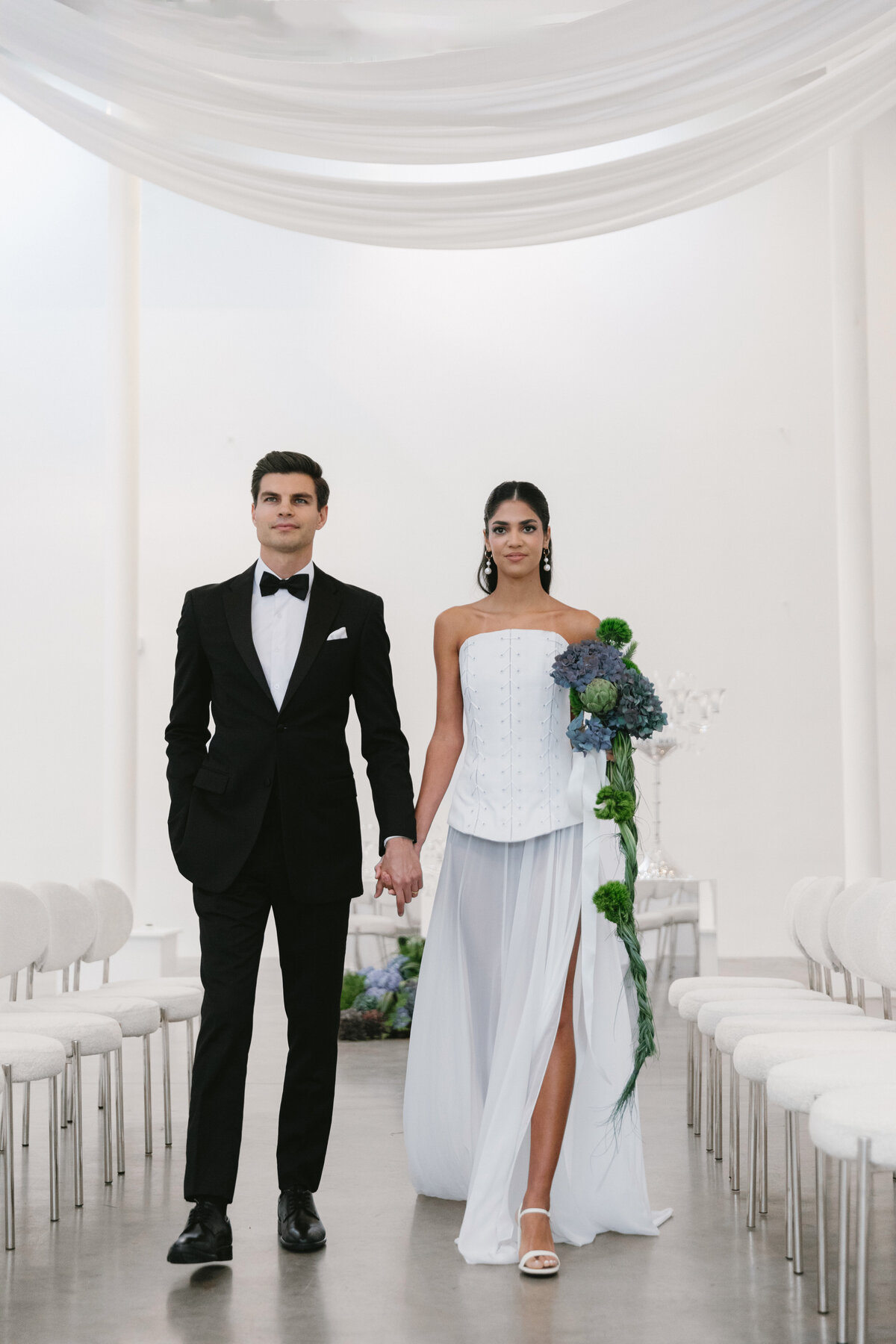 Bride and groom walking hand-in-hand down a modern white aisle with flowing drapery overhead at an editorial New York City wedding shoot.