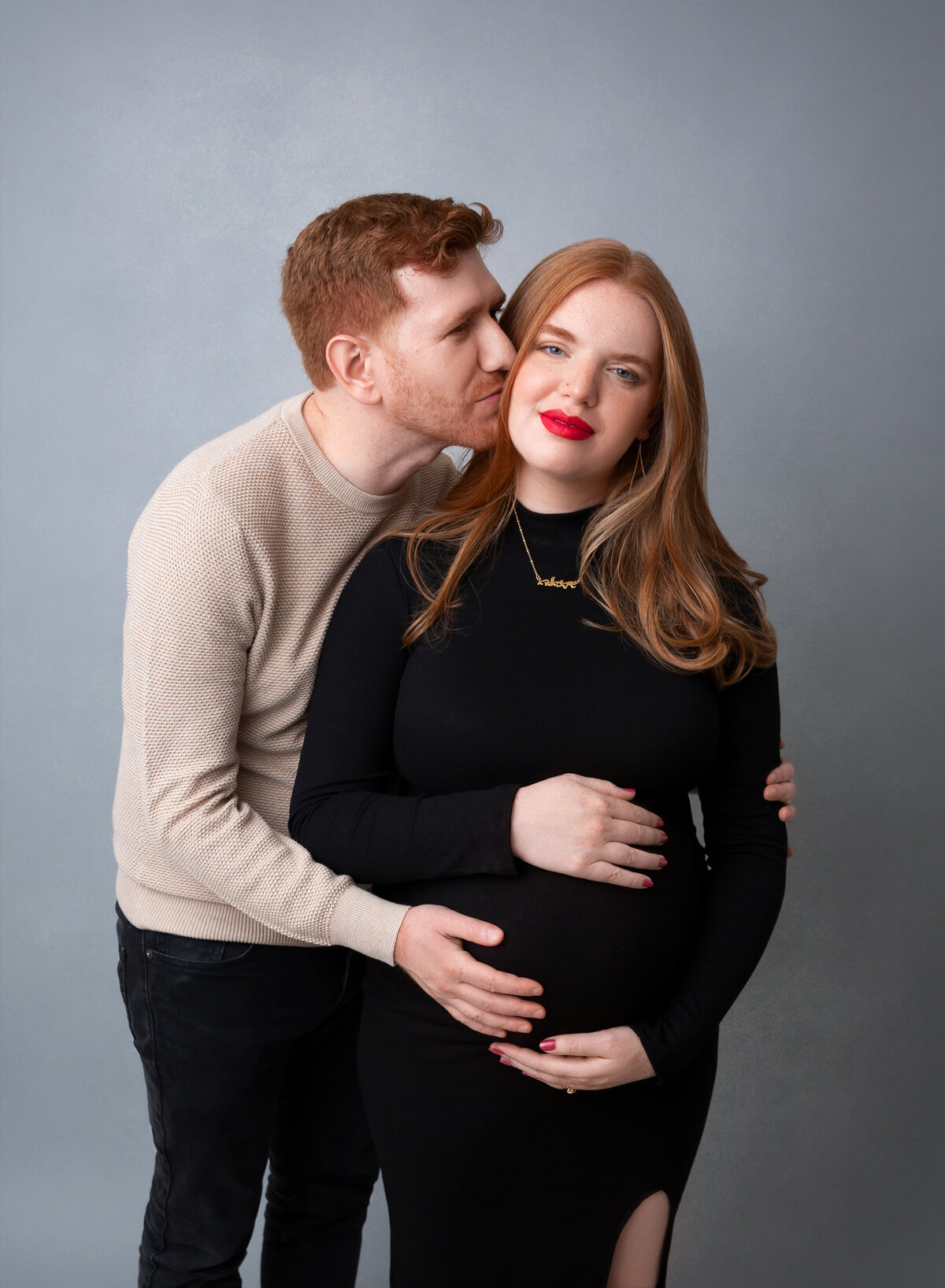 Expectant mom in fitted black dress poses for maternity photoshoot in Brooklyn, NY. Mom has one hand above and below her bump. Expectant dad stands behind mom kissing her cheek.
