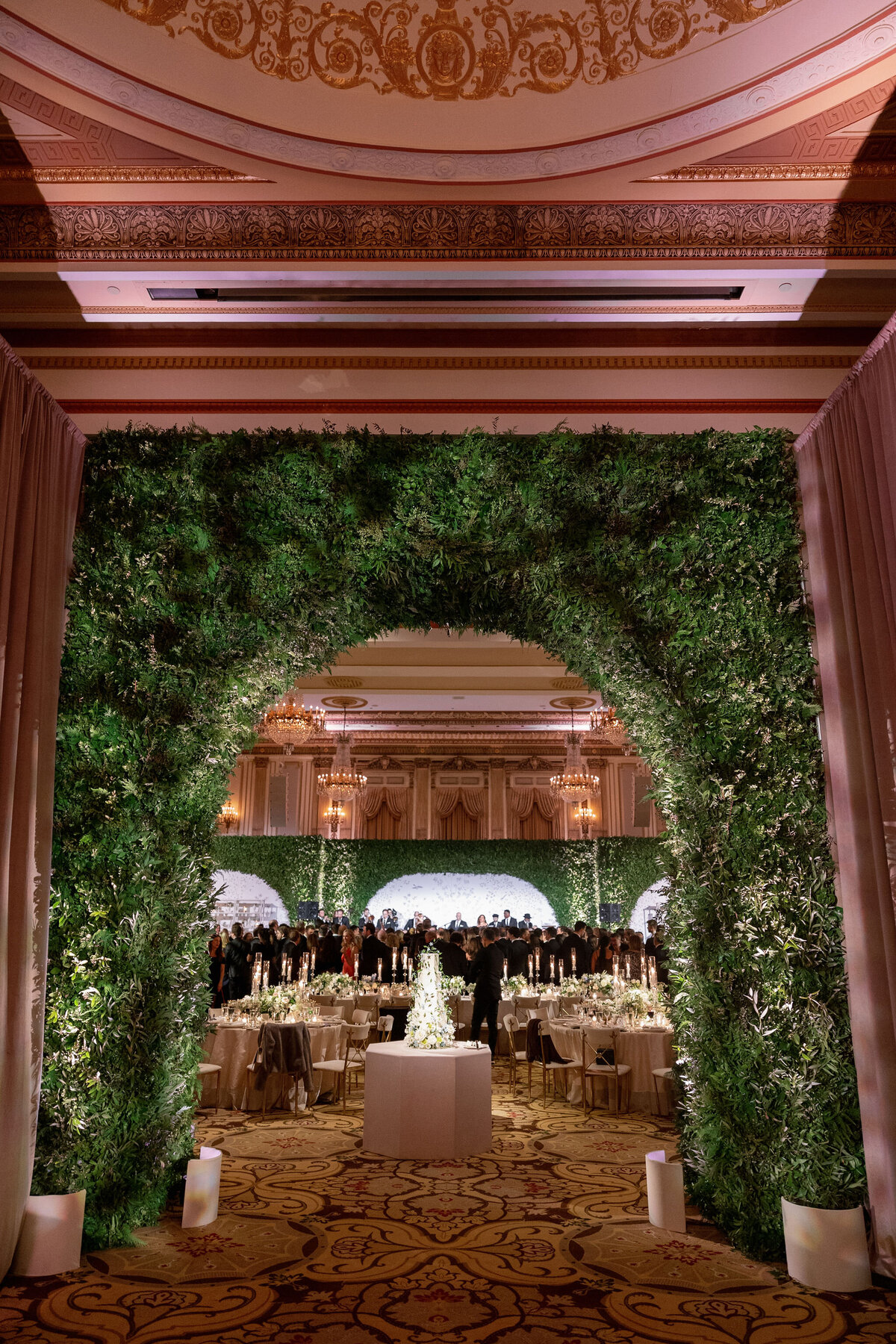 Grand wedding hall entrance framed by a lush green archway, leading to elegantly set tables.