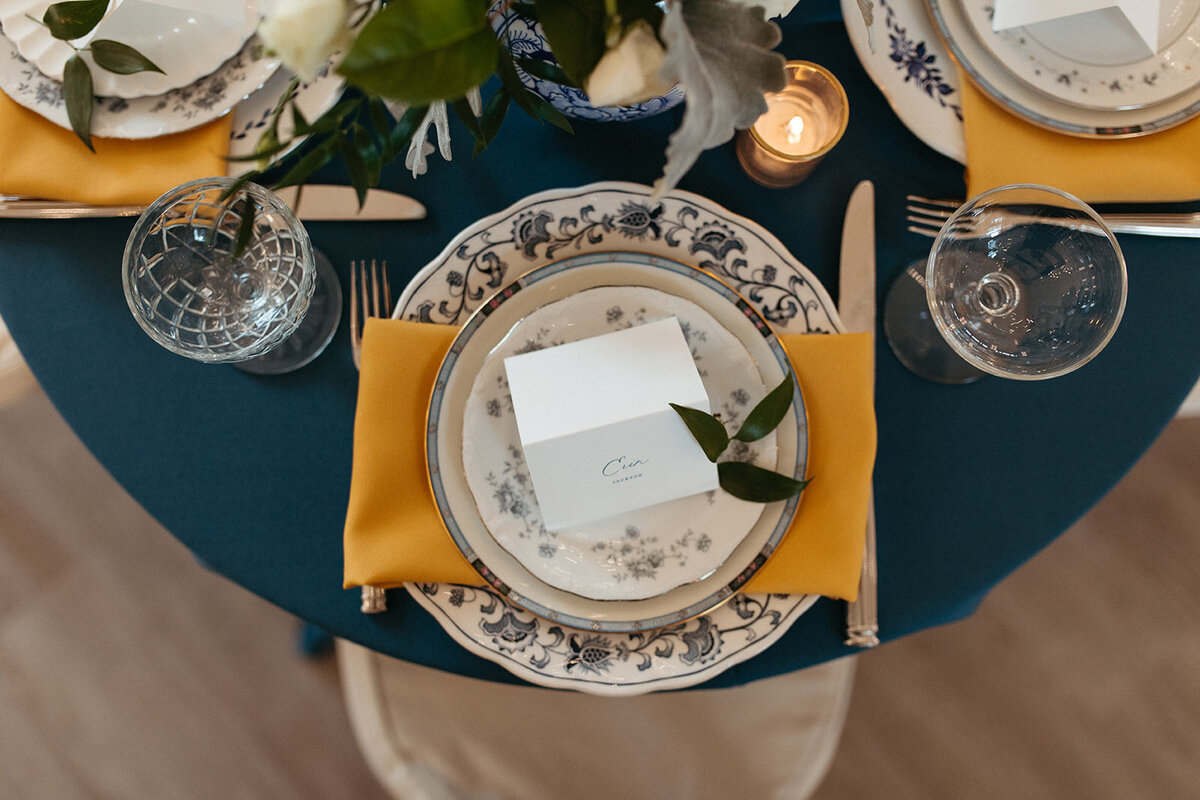 White placecard with gray font atop blue and white plates and napkin on round table with blue linen and candles.