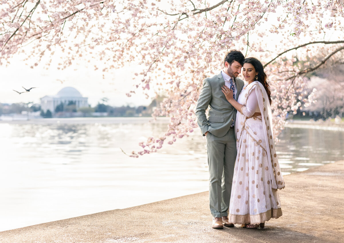 A couple embraces under cherry blossoms by a serene lake. The man is in a light gray suit, and the woman is in a white and gold sari. The Jefferson Memorial is visible in the background.