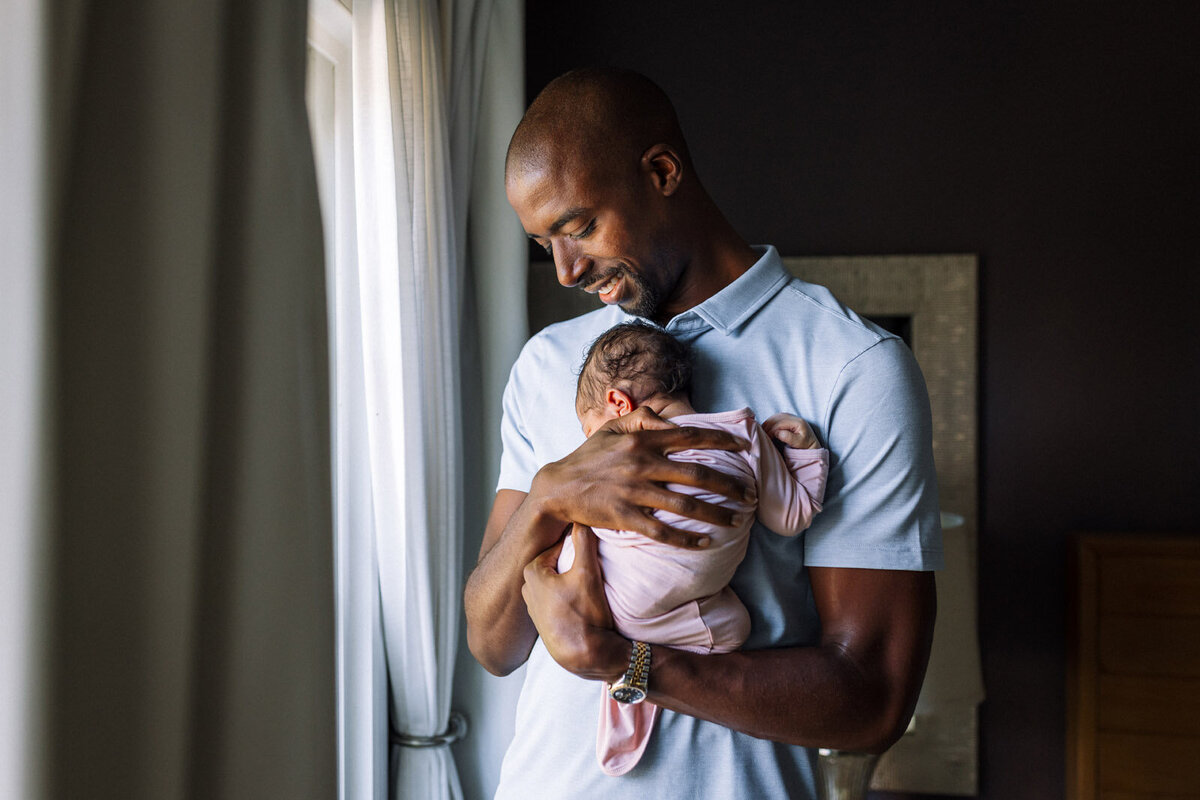 dad holding newborn daughter next to window looking down and smiling at her
