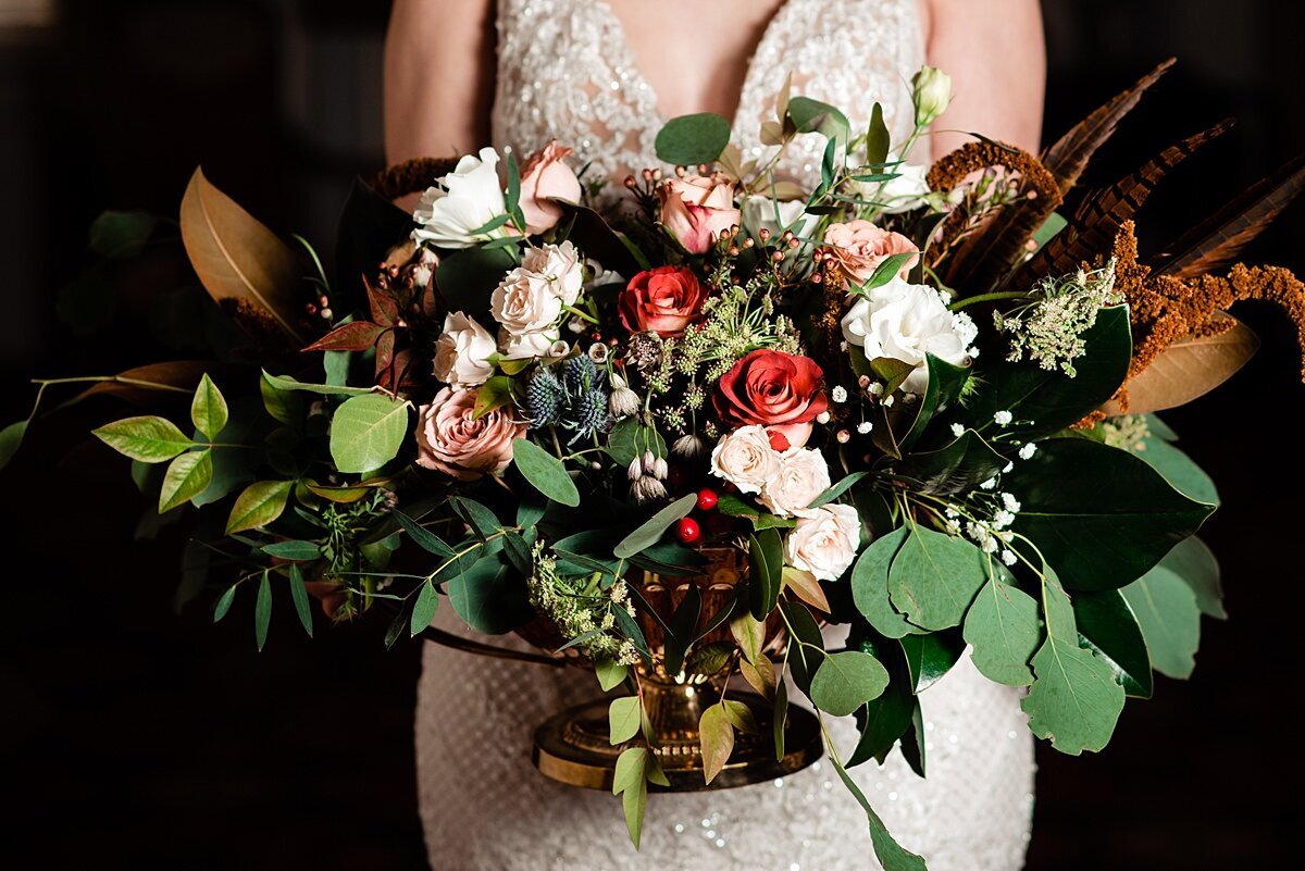 A horizontal bridal bouquet with assorted greenery, magnolia leaves, white roses, blush roses, orange roses, red roses, burgundy dahlias, berries and veronica.