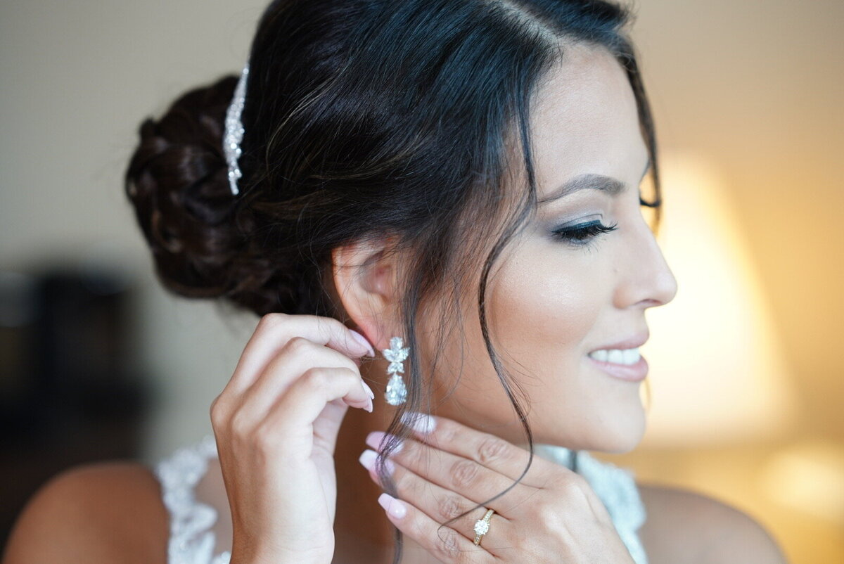 A bride carefully puts on her earrings, focusing on the final details of her bridal look. This image captures a close-up view of her delicate accessories and the elegance of her preparation, showcasing the sophisticated and personal moments before her wedding.