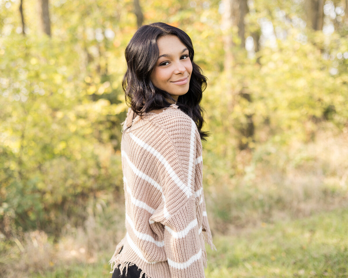 high school female senior, with long black hair, standing in grass with a tree line next to her, standing to the side, looking at the camera smiling softly, wearing black leggings, and light tan and white stripped shirt