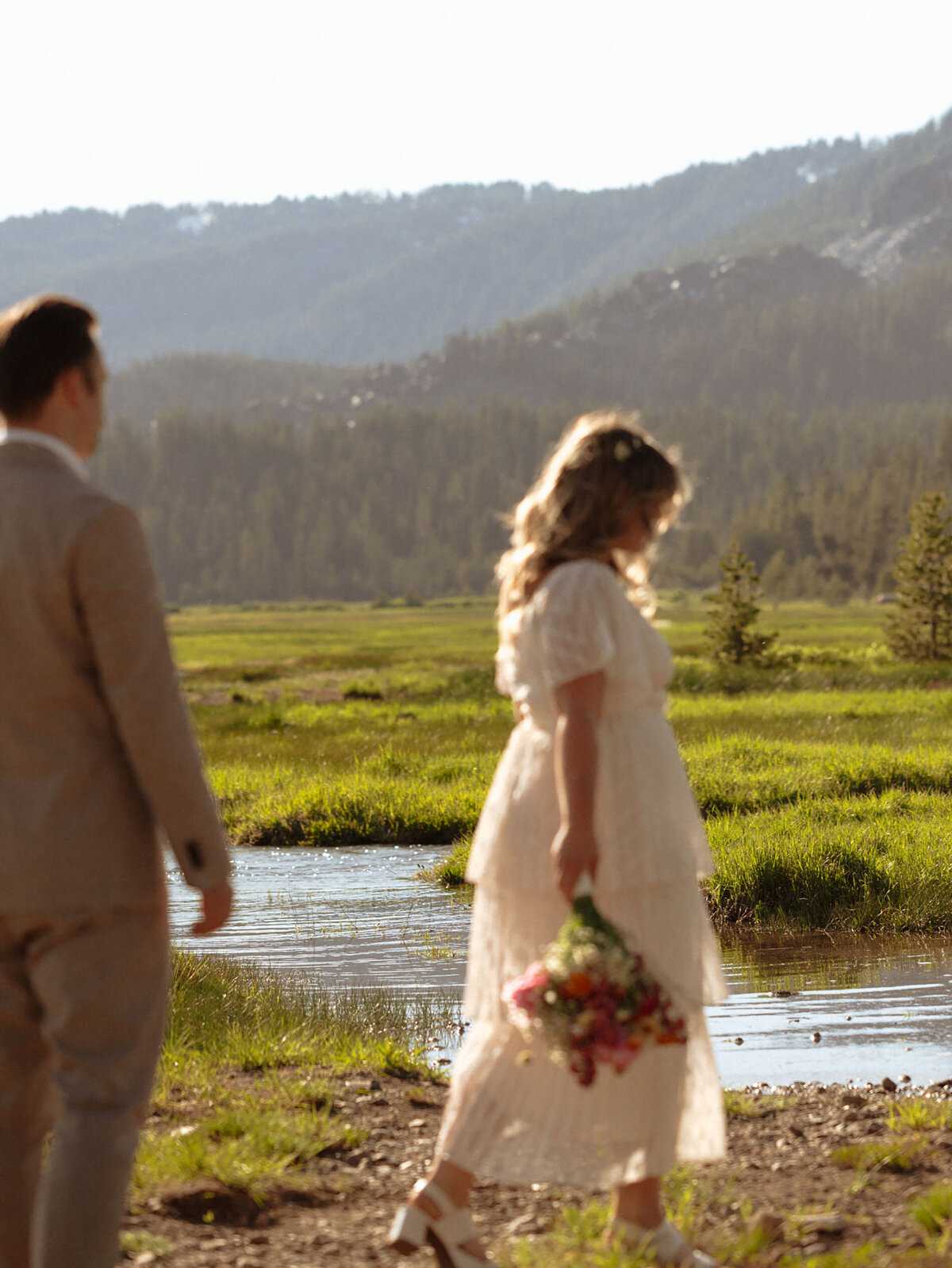 bride walks along lake while holding flowers while the groom follows closely behind