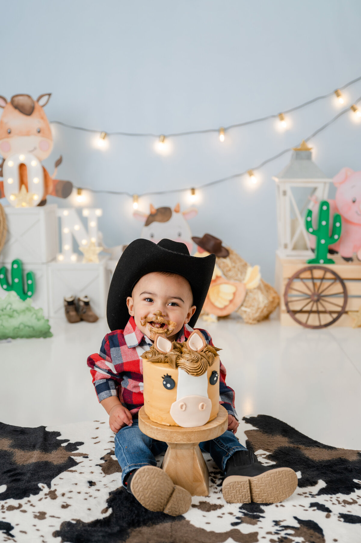 Smiling baby boy in a black cowboy hat sits on a cowhide rug next to a horse-themed cake.
