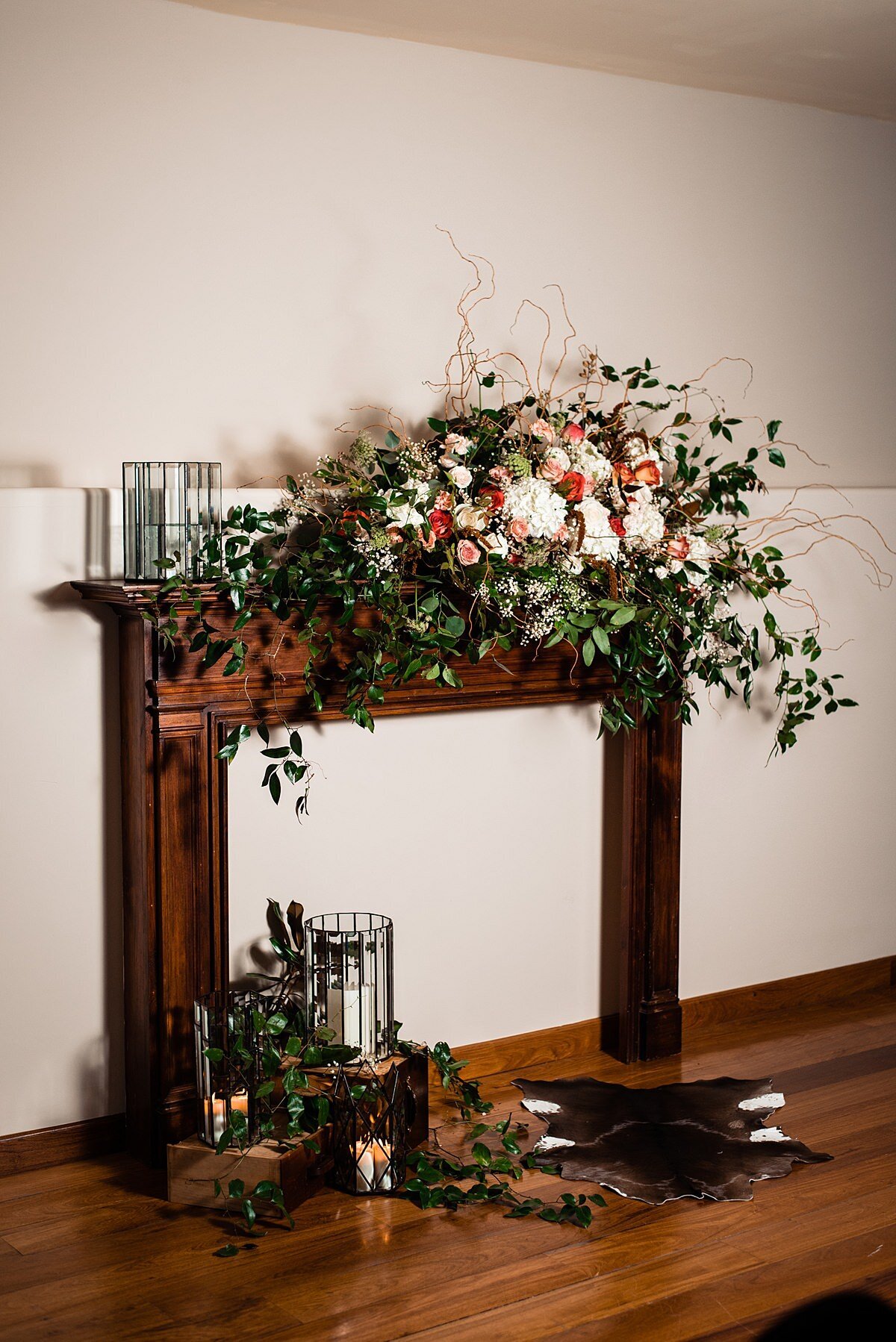 A dark wood mantle against a white wall decorated with beveled glass cylinders holding candles. A cow skin rug lays on the floor in front of the mantle on a dark wood floor. An extra large asymmetrical floral centerpiece sits on the right hand side with greenery vines, white hydrangea, blush roses and burgundy roses.