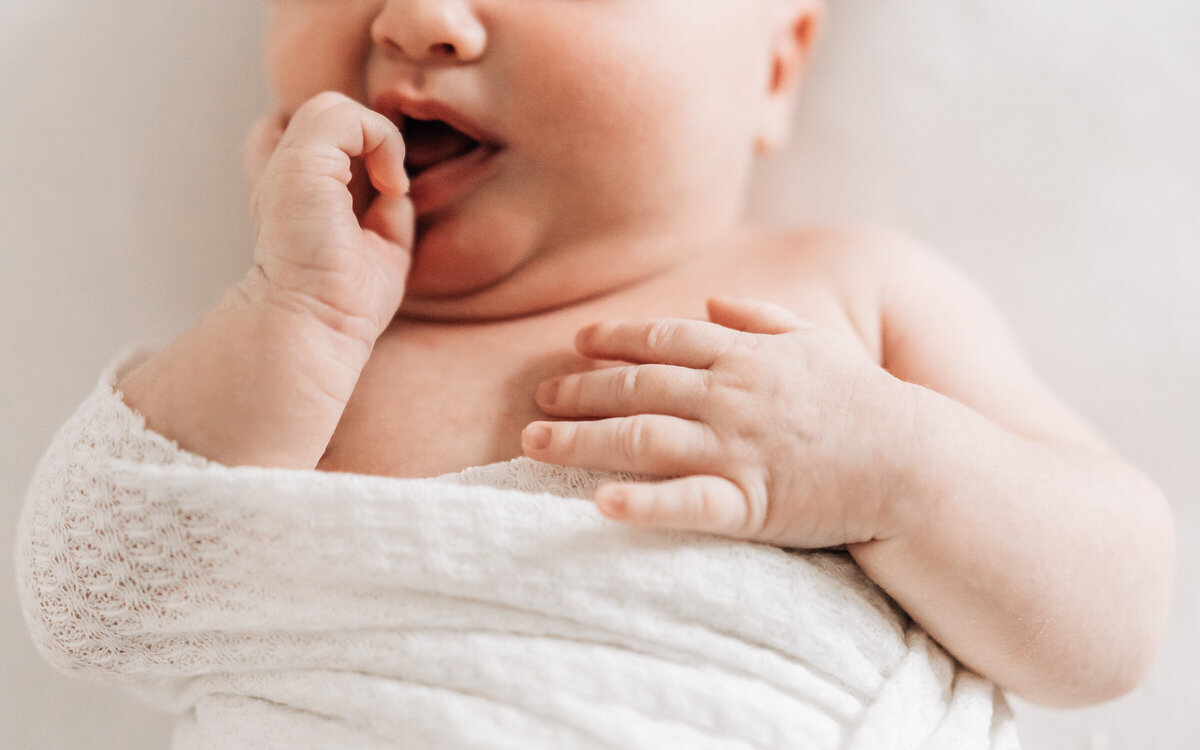 A closely cropped image of a partially swaddled newborn baby with a hand in her mouth during an in home newborn session by Minneapolis photographer, Kate Simpson.