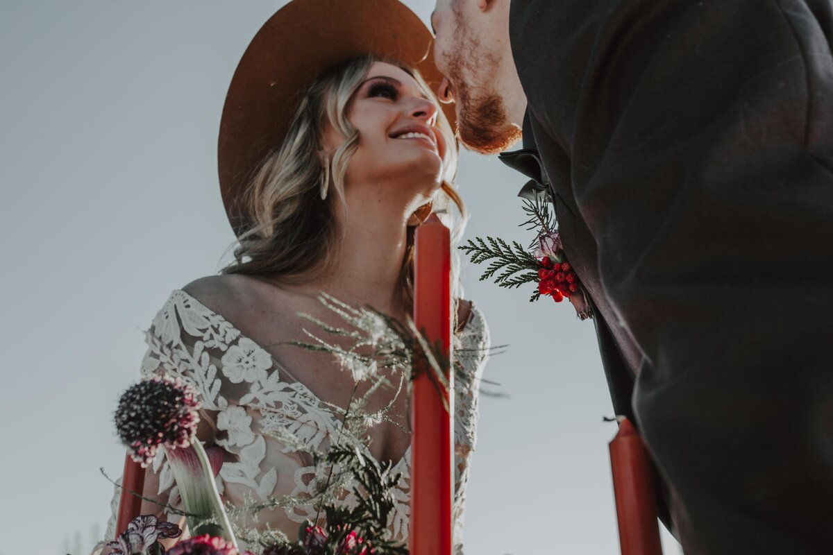 boho mountain wedding with bride in a lace long sleeve wedding dress and a trendy hat leaning over a table with orange candles and florals to kiss her groom