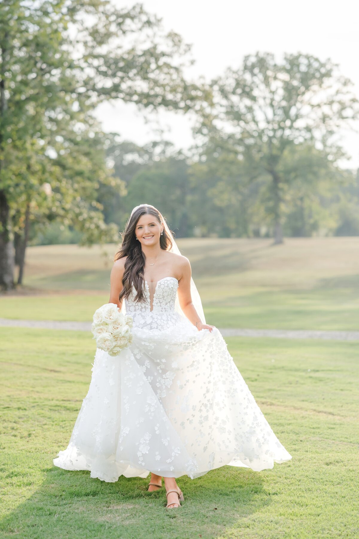 A happy bride holds her lace dress while walking in a manicured lawn at sunset