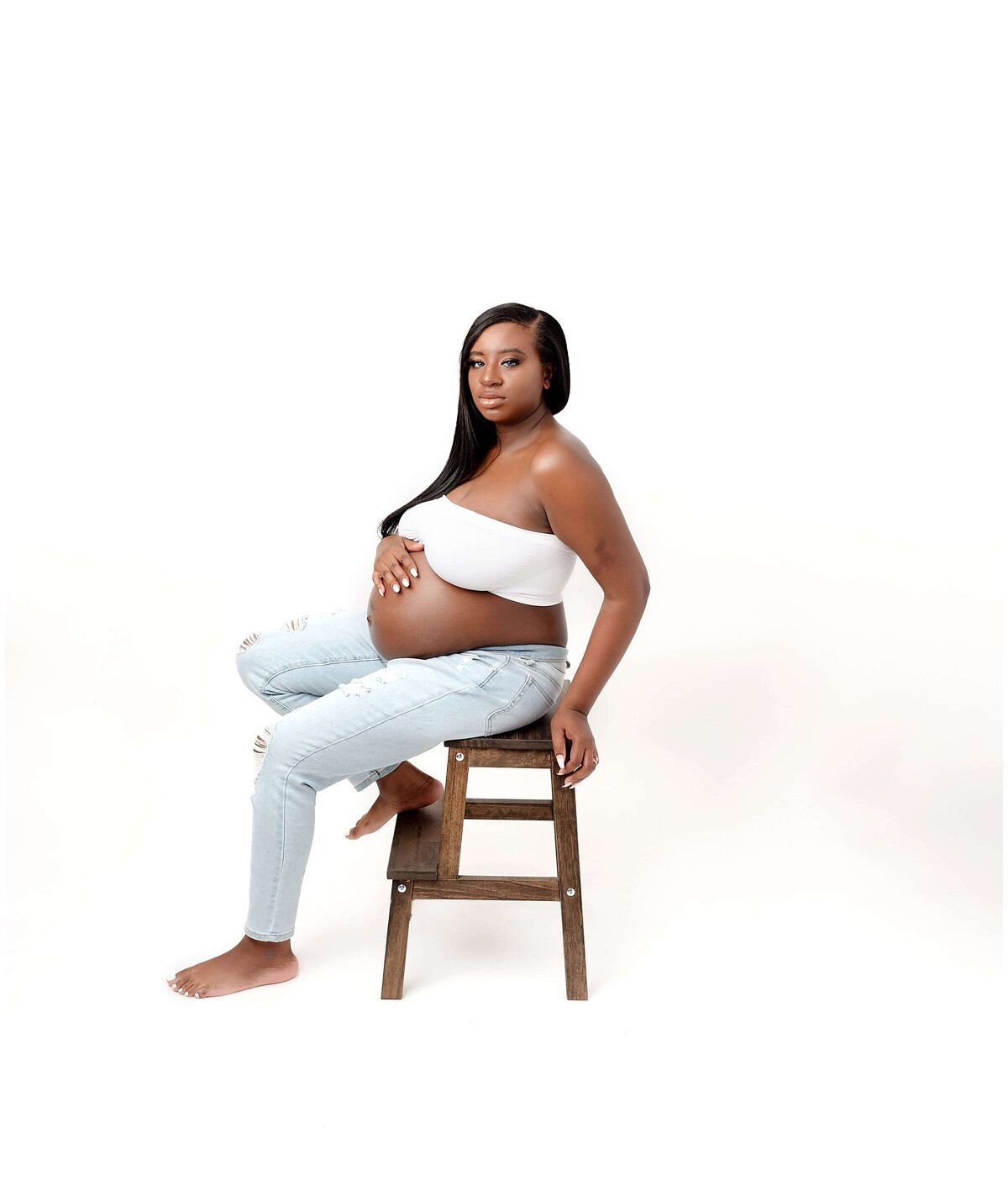 A pregnant woman sitting on a stool, wearing jeans and a tube top outfit. She rests her hands on her baby bump, showcasing the bond between mother and child. The soft lighting highlights her radiant glow, capturing the beauty and serenity of this maternity session.