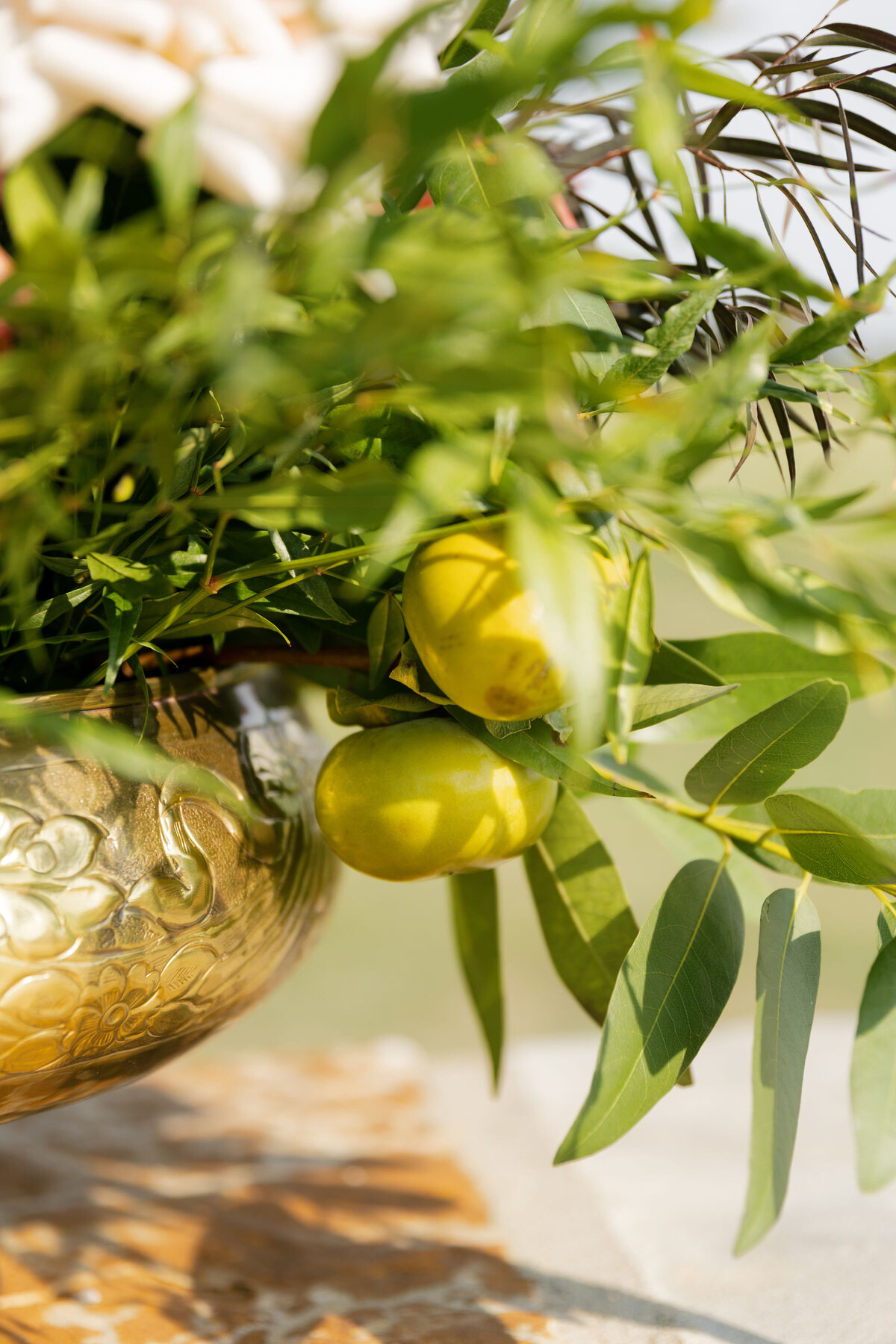 Detail photograph of fresh green fruit nestled among lush greenery inside a textured gold wedding vase, adding organic Mediterranean-style accents to the ceremony décor.