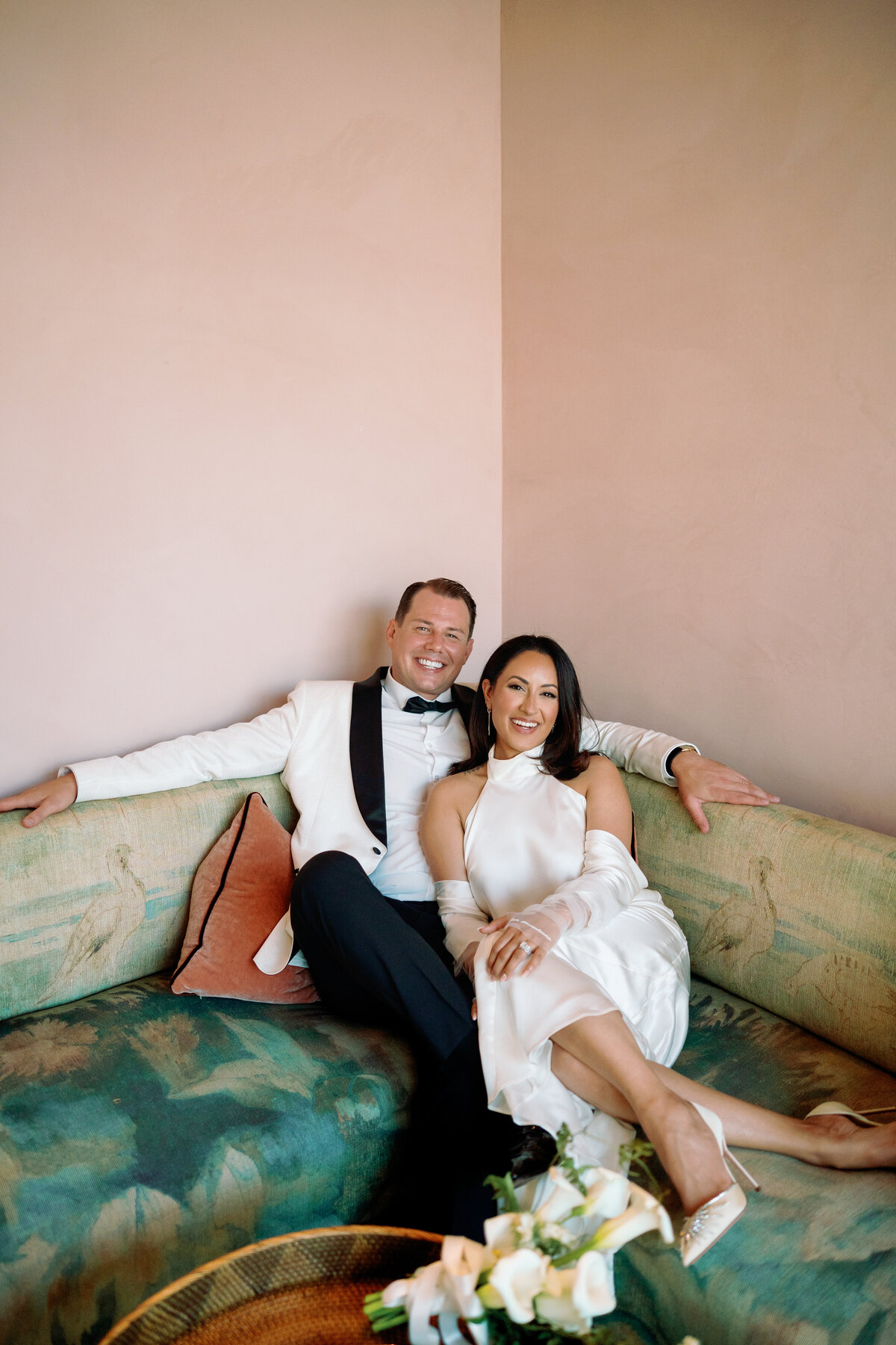 Couple lounges together on a pastel vintage sofa, laughing and enjoying a quiet moment on their wedding day.