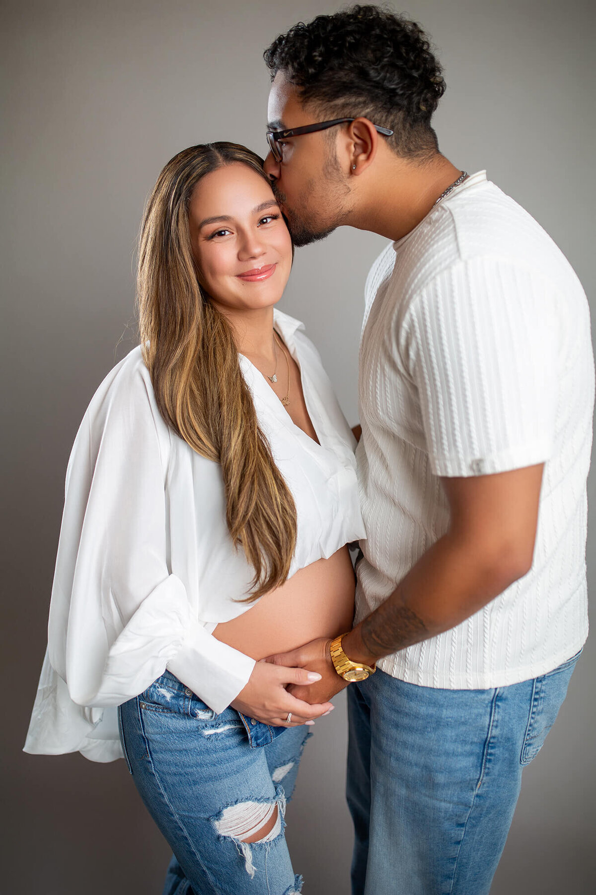 Man is kissing woman with long brown hair while holding pregnant belly in the studio maternity session 