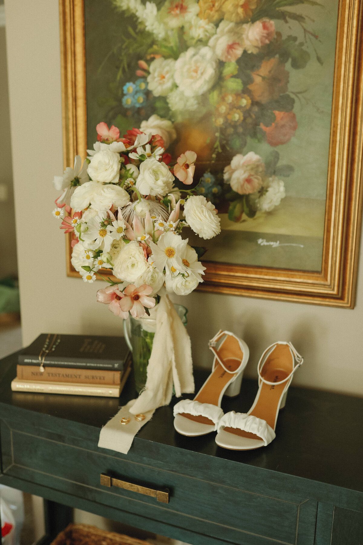 brides wedding shoes and flowers sitting on green dresser with flower painting  in the back round