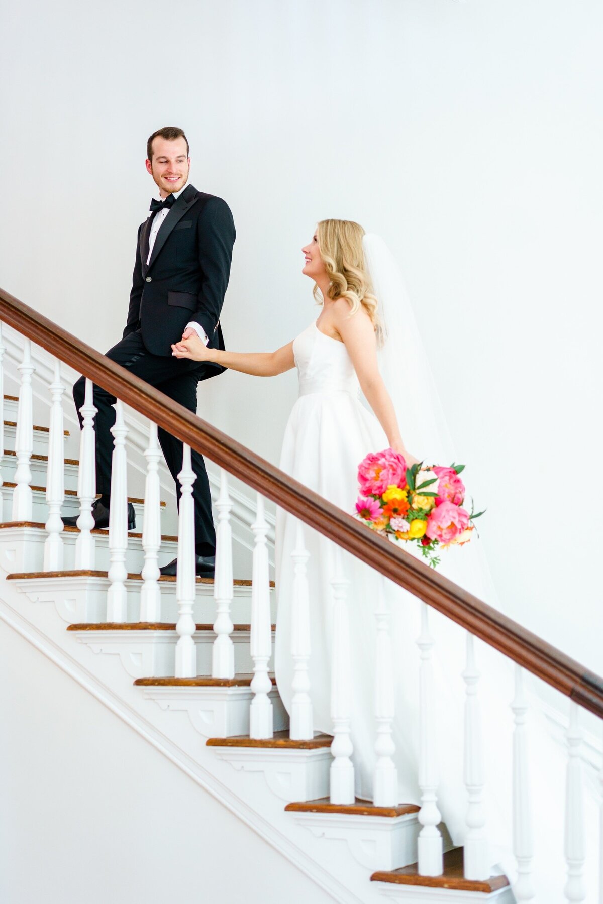 A groom leads his bride up a historic white staircase at their Raleigh wedding venue.
