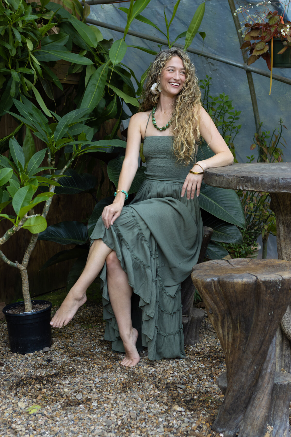 Dr. Mamie Burruss, ND, LAc sitting casually at a wooden table outside wearing a sage green dress and surrounded by foliage