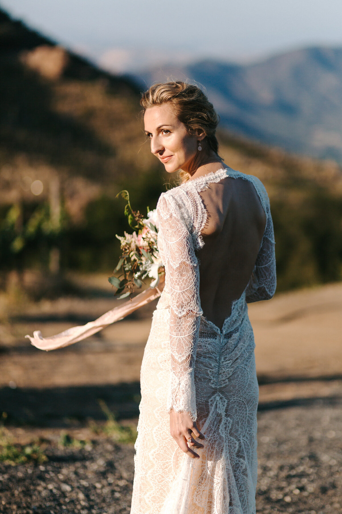 A colorful portrait of Claressinka on her wedding day at Saddlerock Ranch in Malibu, California. Claressinka has a lace dress on with long sleeves and an open back. Her body is facing the Malibu hills behind her, but she is turning her head to the left of the camera. The ribbon from her floral bouquet is blowing in the wind. Wedding photography by Stacie McChesney/Vitae Weddings.