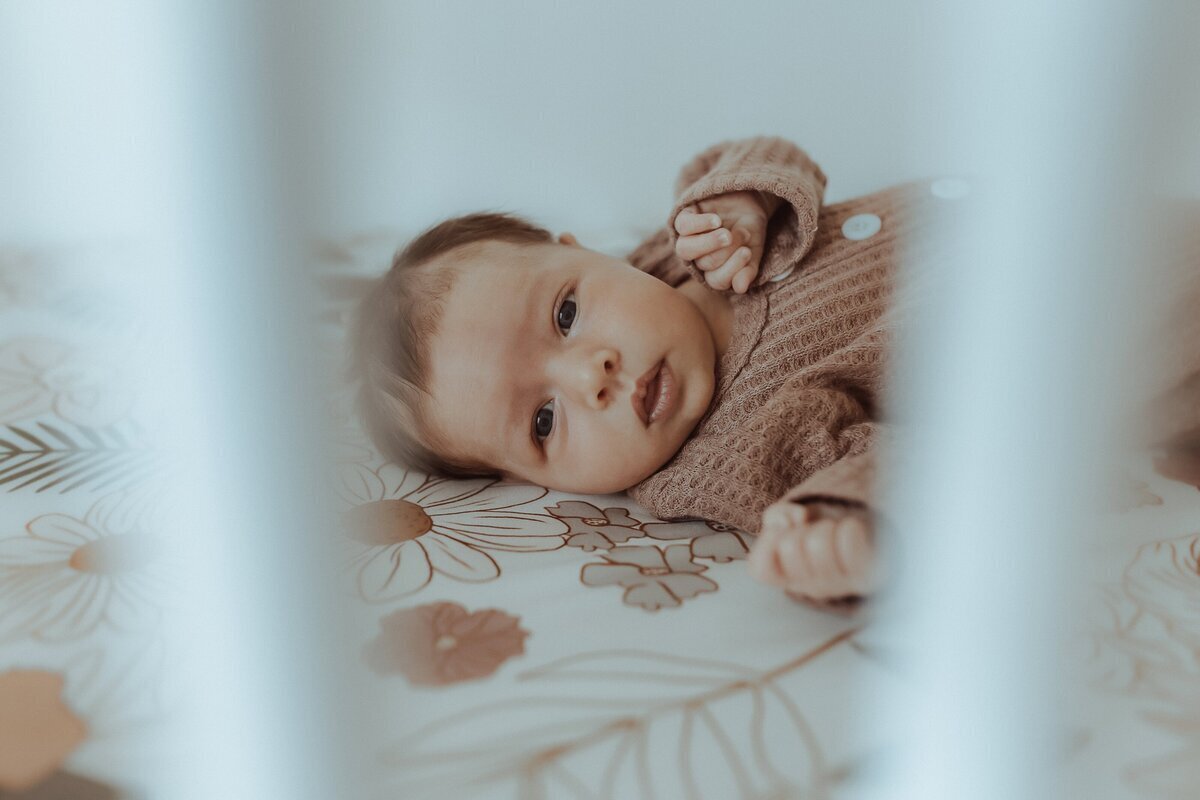 Baby lying on a floral-patterned blanket, viewed through the slats of a crib, wearing a textured brown outfit.