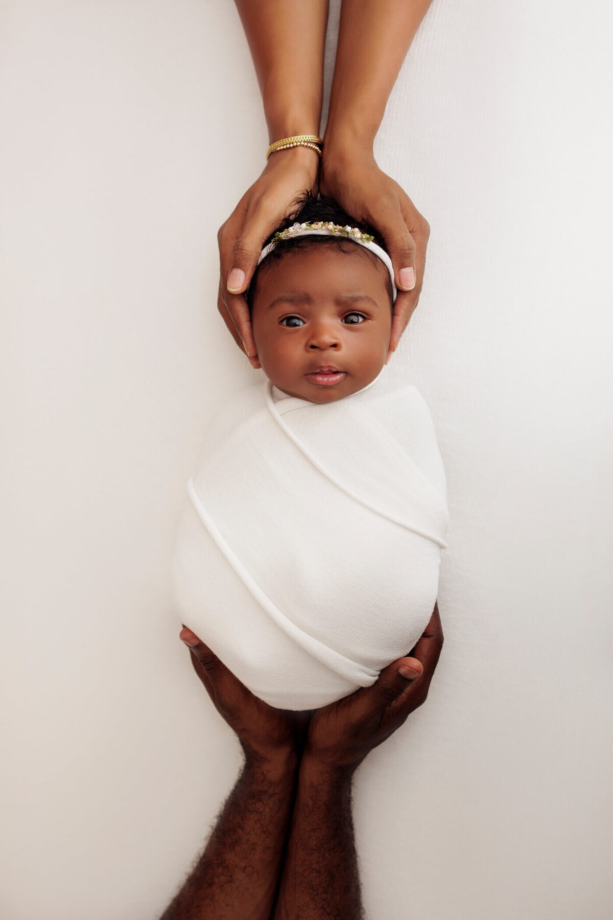 Newborn baby girl wrapped in a white swaddle, held securely by her parents' hands from above and below, looking directly at the camera against a white background.