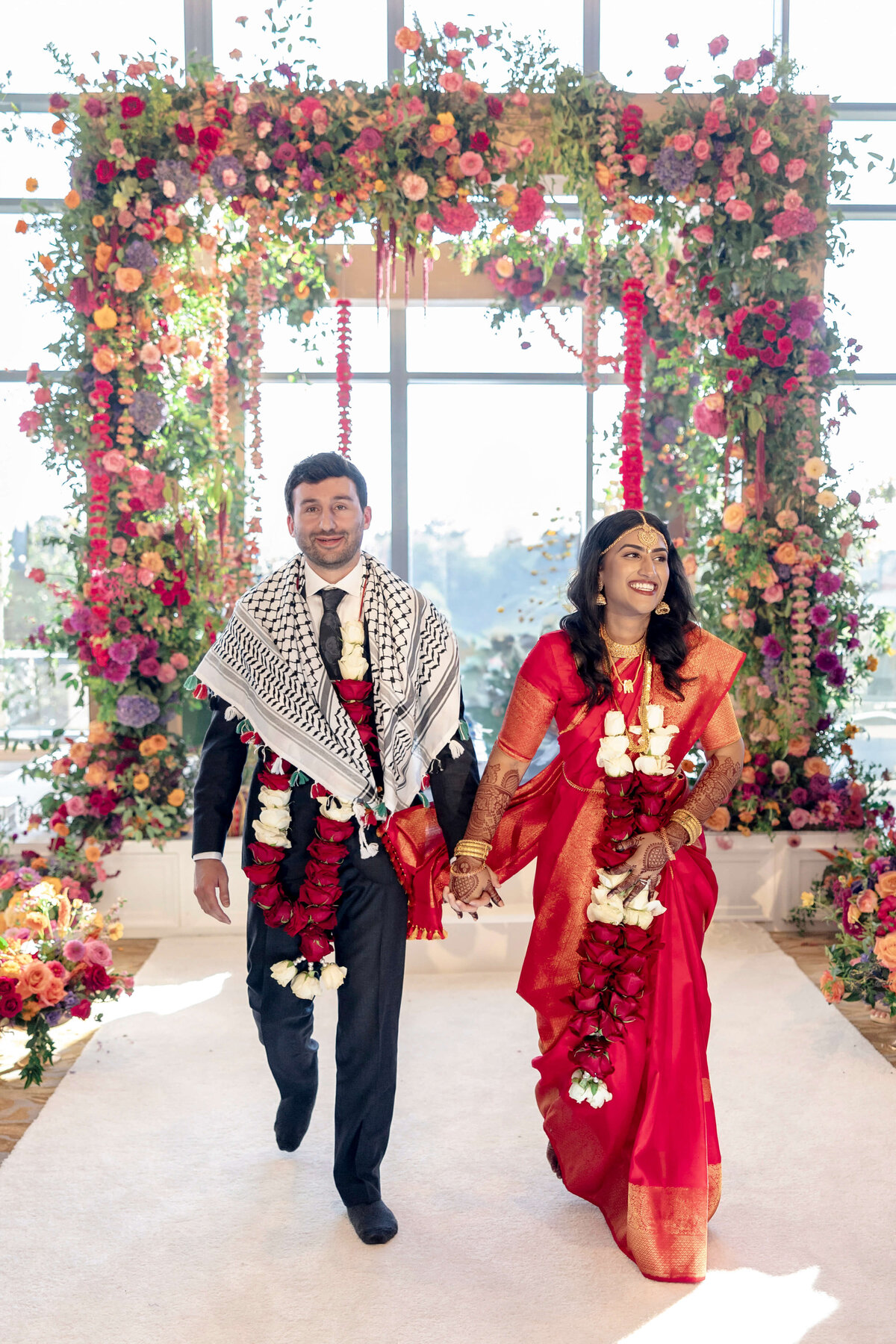 A couple walks hand in hand, smiling, under a vibrant floral arch. The woman wears a red sari and garland, while the man is in a suit with a patterned shawl and garland. Bright flowers surround them, and sunlight filters through large windows in the background.