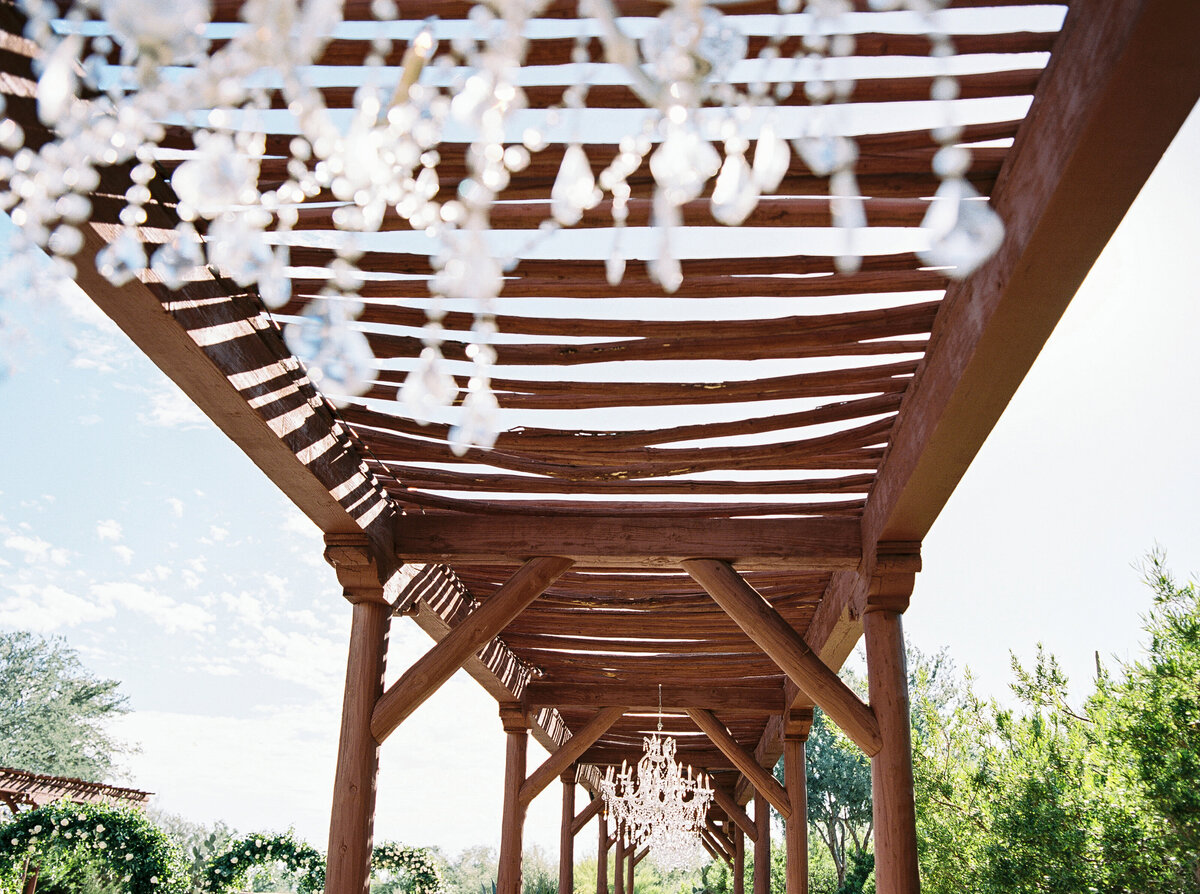 A beautifully designed rustic wedding venue featuring an elegant wooden pergola adorned with crystal chandeliers under an open blue sky.