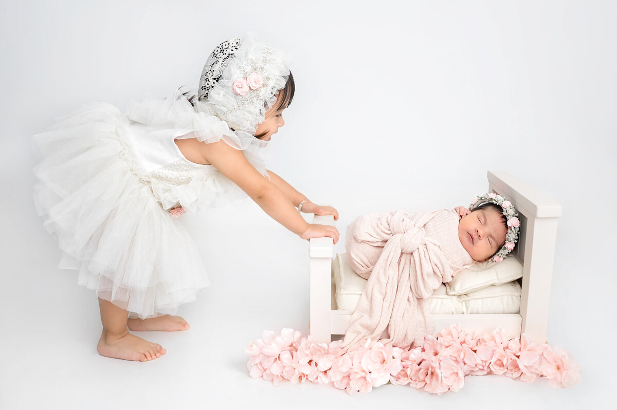 Older child stands over newborn baby who is wrapped in soft pink fabric while laying on a small off white bed. 