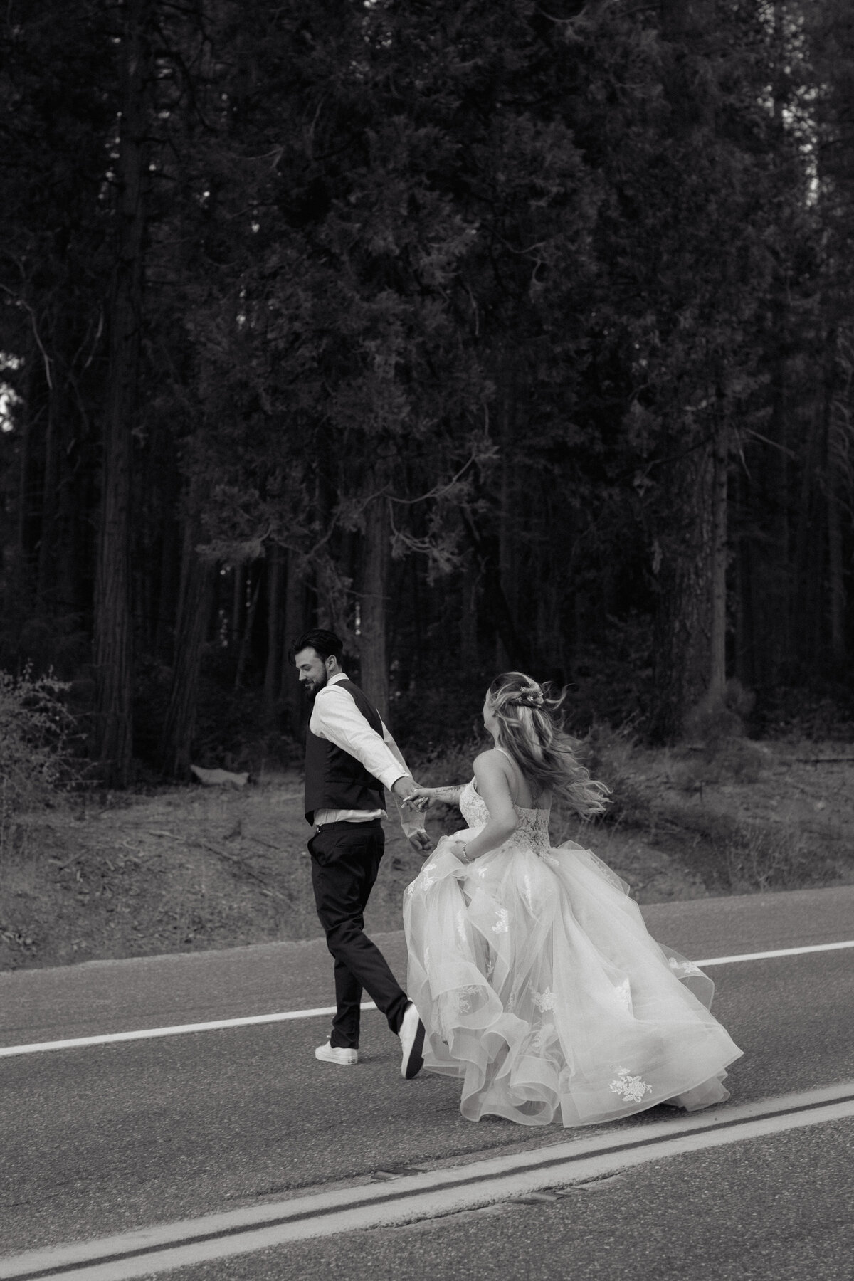 Black and white wedding photo of a bride and groom running hand in hand down a road, blending documentary wedding photography with an adventurous, cinematic feel.
