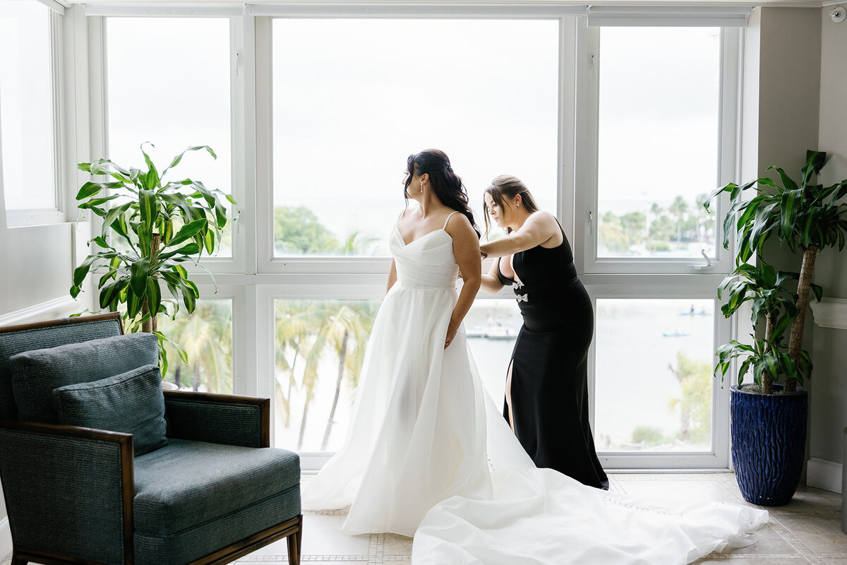 The bride standing by a window as her bridesmaid helps adjust her gown, with a view of Ocean Reef in Key Largo in the background, captured by Claudia Amalia Photography during Ashley and Cameron's micro wedding.