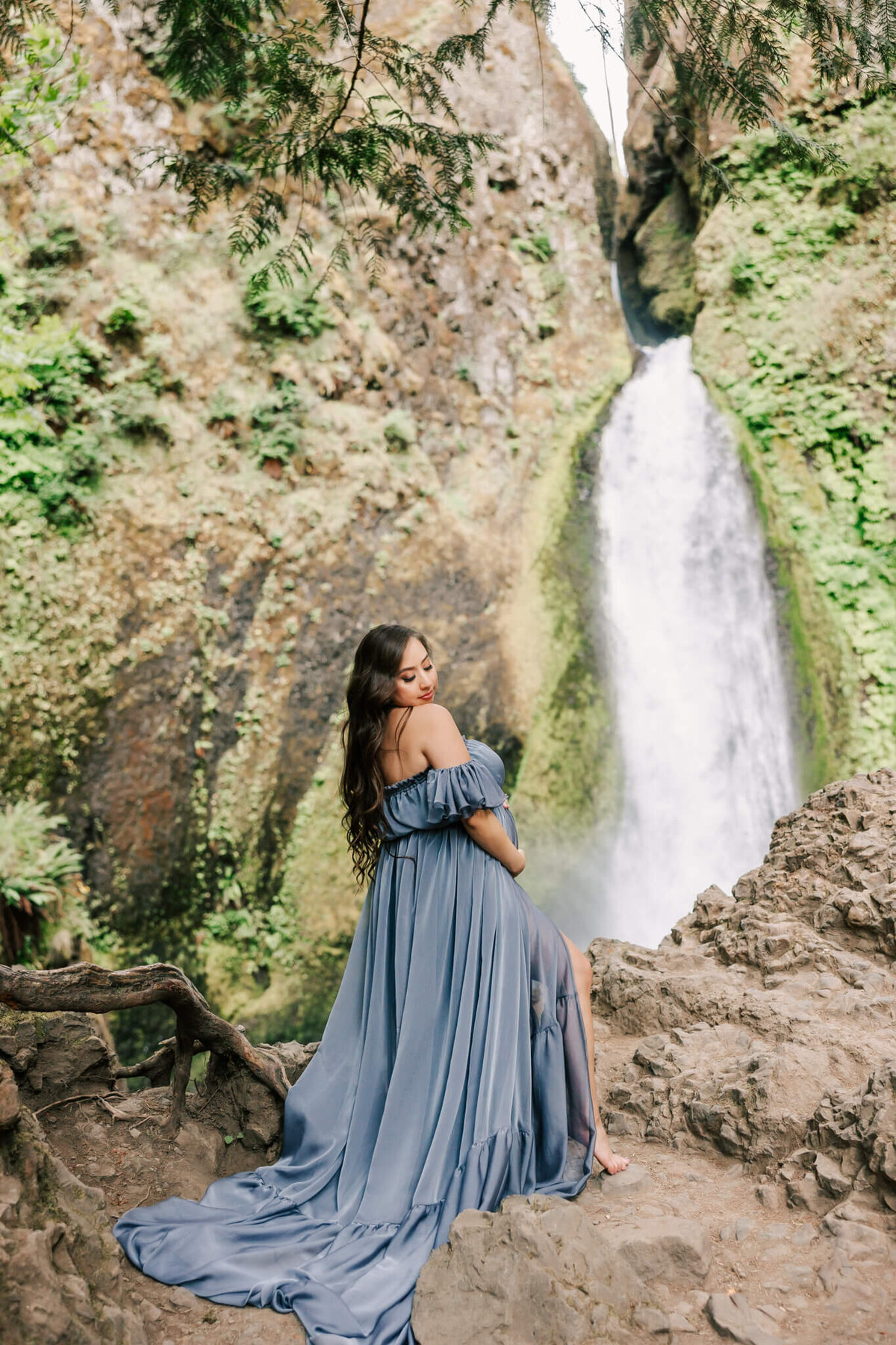 Woman in blue dress standing in front of waterfall in the columbia river gorge. She is looking over her shoulder and has her hands on her belly.