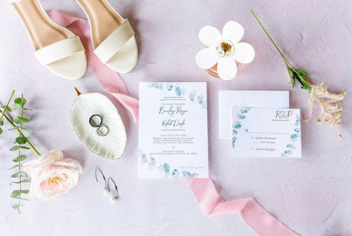 Close-up wedding invitation with rings, perfume, flowers, and shoes.