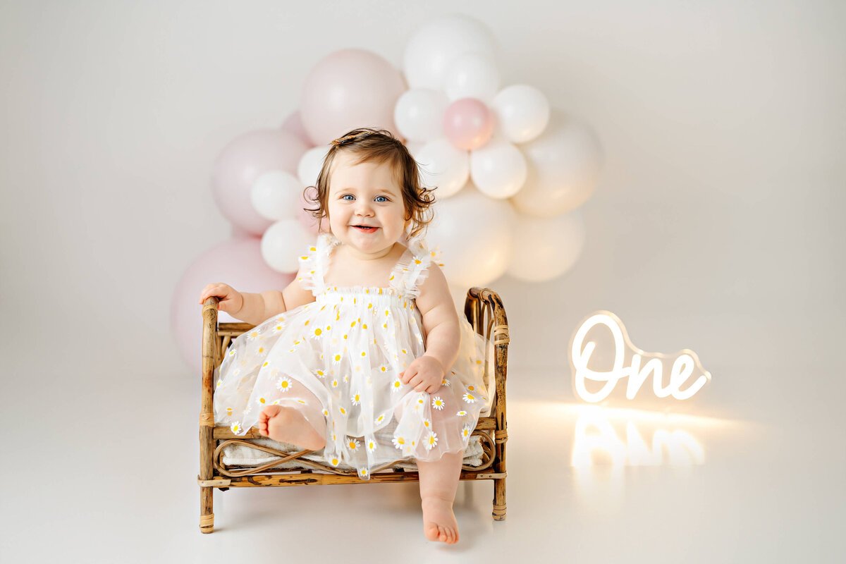 A smiling baby in a white dress with yellow flowers sits on a small wicker bench. Behind her, there are white and pink balloons, and a glowing sign that reads "One." The setting is bright and minimalistic.