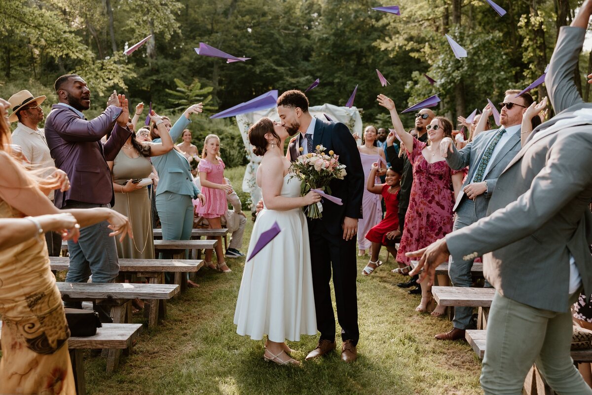 Bride and groom share a kiss during their outdoor wedding ceremony exit as guests toss purple paper airplanes in celebration.