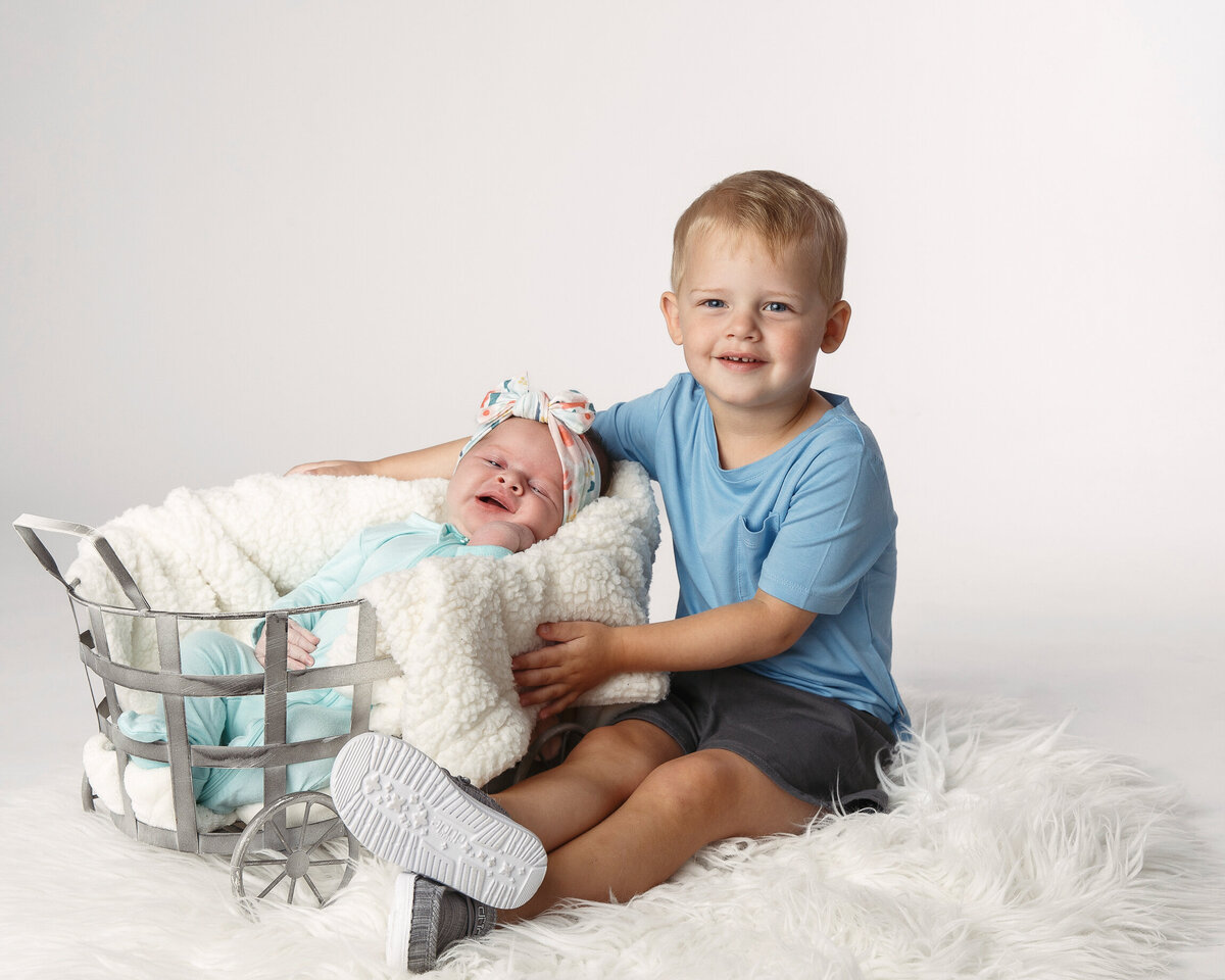 Three year old boy wearing a blue shirt posed next to a basket with his newborn baby sister