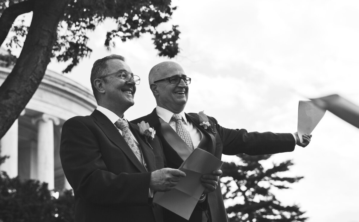 Two people in suits smiling together while one holds a piece of paper