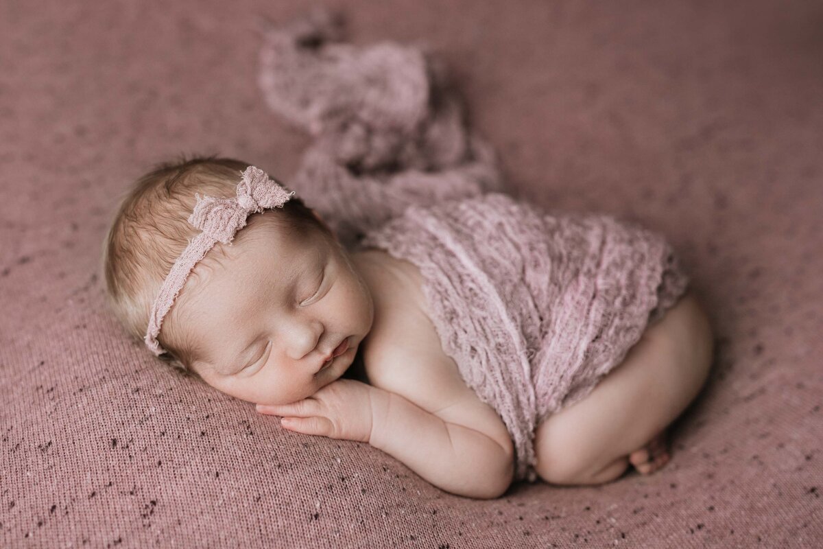 Studio newborn photography - baby sleeping on her side curled up. She has a mauve swaddle draped around her belly with a pale pink bow headband.
