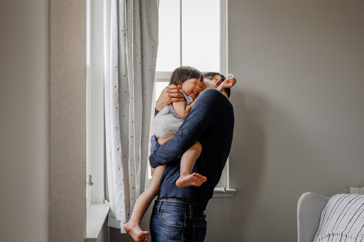 dad holding 4-year-old daughter close and making her laugh while standing next to two windows at home