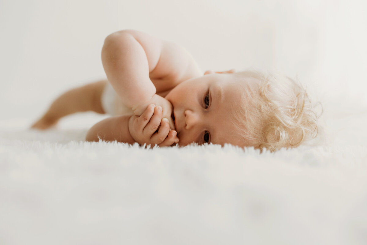 Six month old baby boy laying on his side on a white rug with a white background as he nibbles on his fingers during his Denver Mini milestone session. 