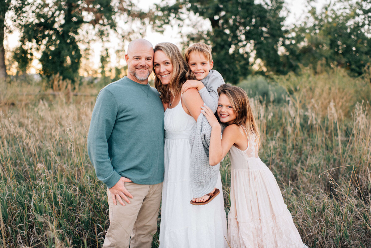 family of four with older school age kids standing in a field at sunset and smiling at the camera