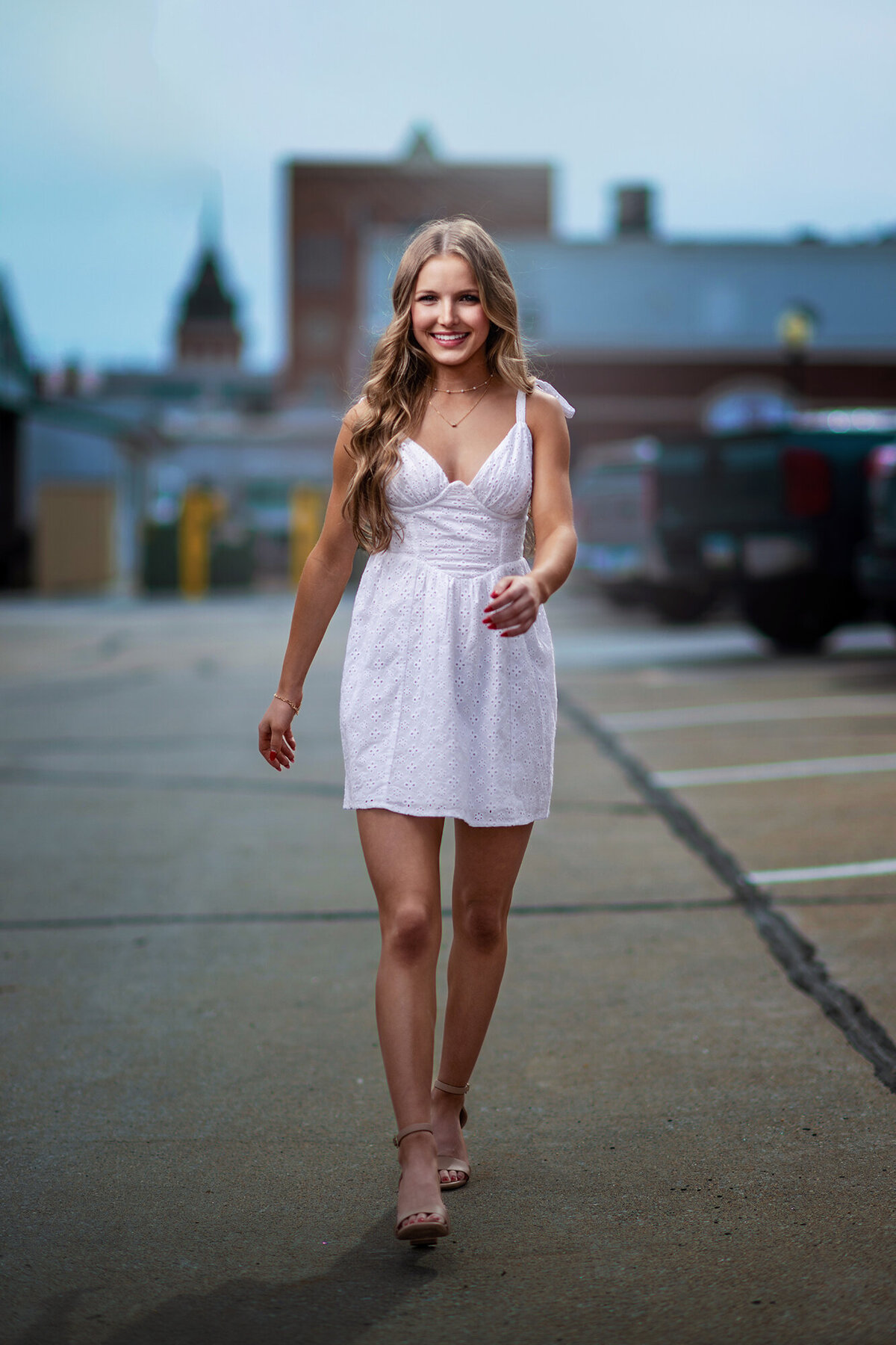 A high school senior in a white dress walks through a parking lot in downtown