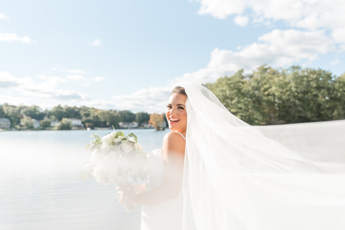 Bride standing on dock by lake with long white veil blowing around her on bright sunny day.